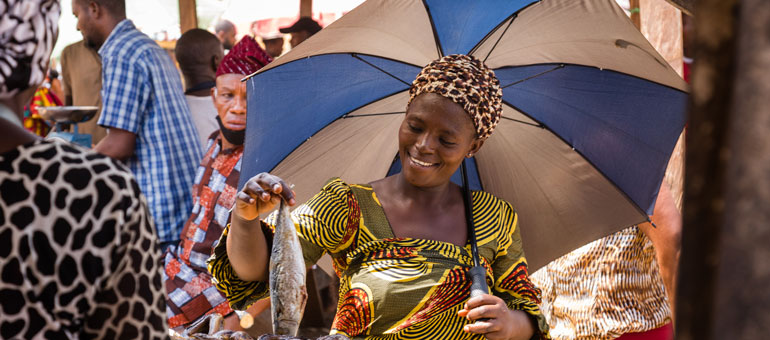 A woman wearing a bright yellow dress and umbrella holding up some dry fish