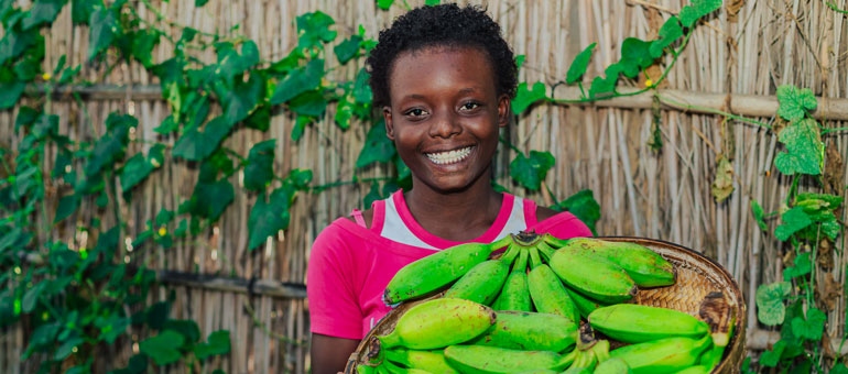 Young girl wearing a pink dress and smiling to the camera while holding a basket of bananas