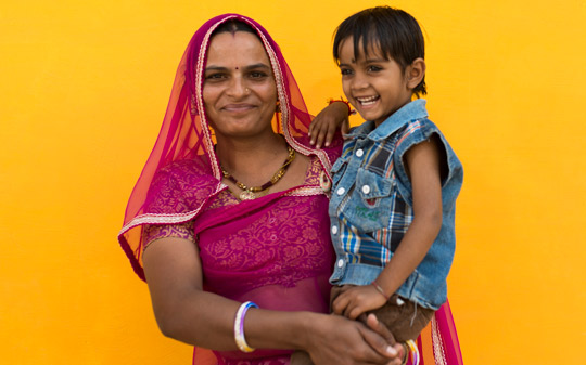 Woman wearing a purple dress and holding her boy against a yellow background in India