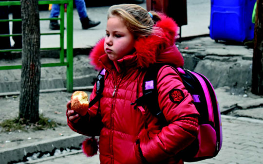 Little girl wearing a red jacket, hair in a ponytail eating croissant in the streets