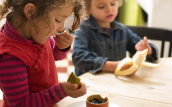 Two children sitting down at the table and eating food with hans and spoons