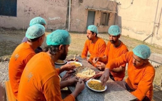 A group of men in orange uniforms seated at a table at work, engaged in discussion while eating