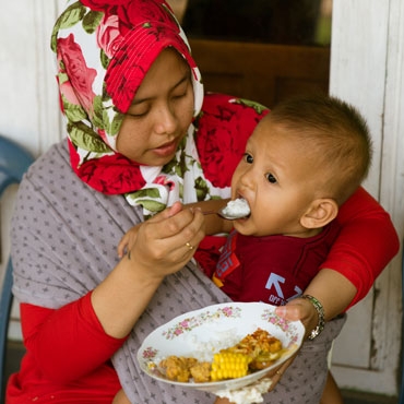 A mother feeding the child with rice and chicken wearing a floral scarf