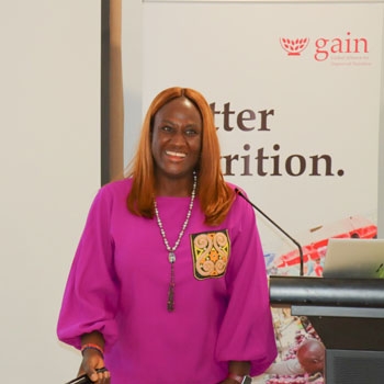 Joyce smiling and wearing a pink blouse in front of the GAIN banner
