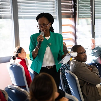 Prisca standing up and talking in the microphone in a room filled with people Prisca standing up and talking in the microphone in a room filled with people